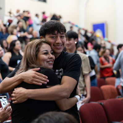 A Student looking into the campera while hugging a family member. They are in auditorium setting. 
